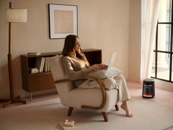 A DREO heater glowing softly next to a window in a bright living room, where a woman works on a laptop in a cozy armchair, bathed in warm sunlight.