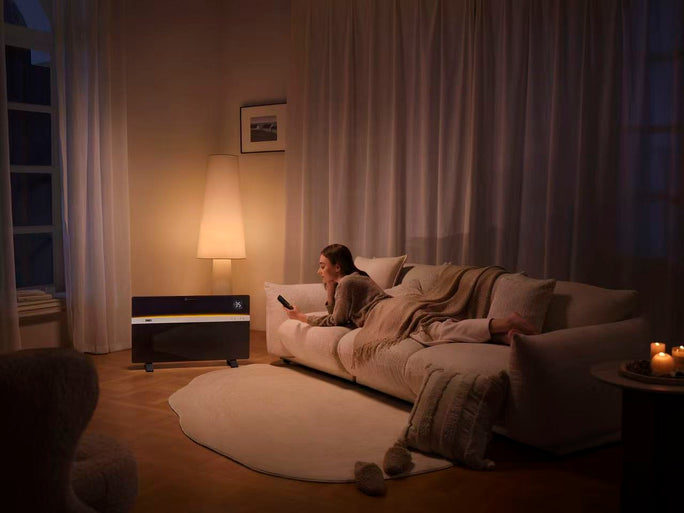 A DREO Convection Heater 711 glowing warmly in a dimly lit living room at night, where a woman lies on a cozy sofa watching TV with a remote control, surrounded by soft lighting from a floor lamp and candles.