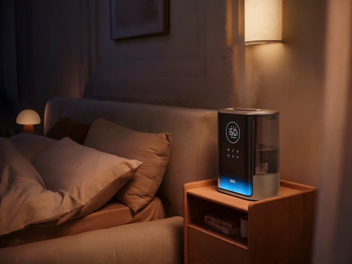 A DREO humidifier sits on a bedroom nightstand at night, displaying 60% humidity with a soft blue ambient glow beside a plush bed.