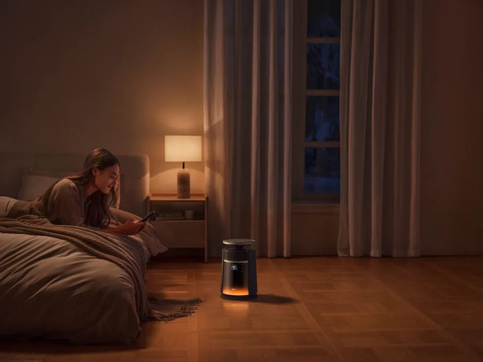A woman lies on her bed scrolling through her phone while a sleek cylindrical DREO heater stands on the wooden floor nearby, casting a gentle, comforting warmth throughout the space.